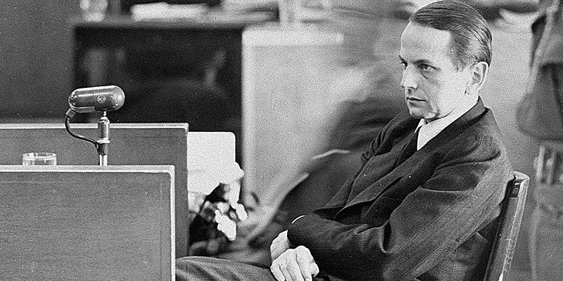 Former SS-Gruppenführer Otto Ohlendorf (Einsatzgruppen D) sits with arms crossed near a microphone in the Nuremberg courtroom, circa 1946–1947.